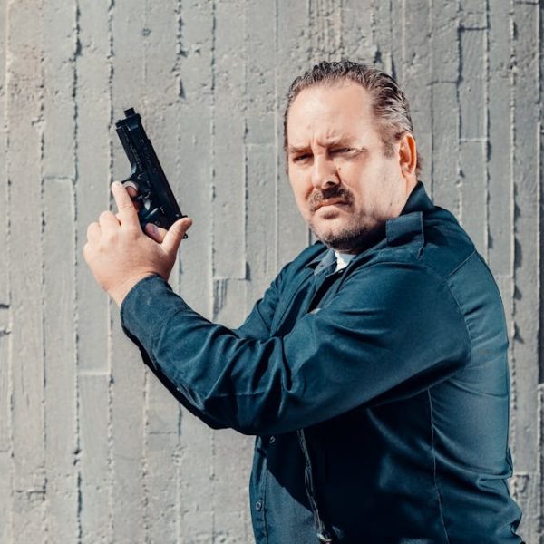 A man in a dark uniform holds a handgun upright beside his face while standing in front of a textured concrete wall.