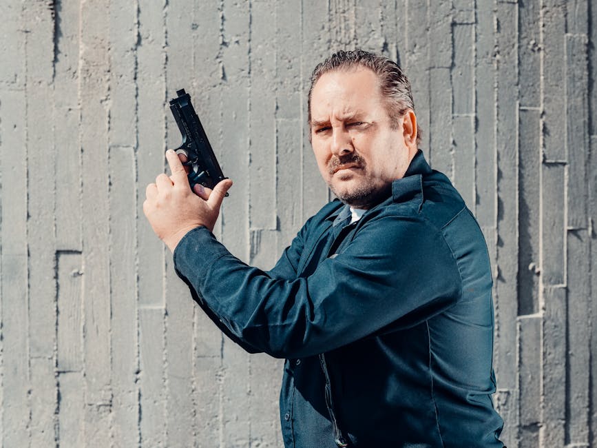 A man in a dark uniform holds a handgun upright beside his face while standing in front of a textured concrete wall.