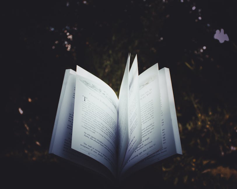 An open book with its pages flipping, set against a dark, blurred background with soft natural light.