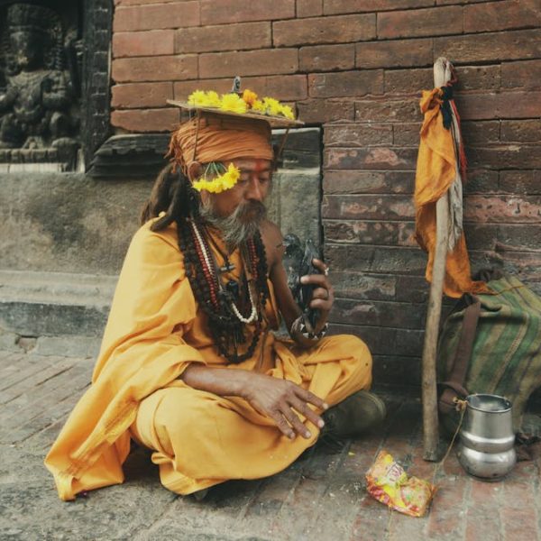 A man dressed in orange robes with flower garlands sits on the ground beside a staff, bag, metal pot, and snack packet, near a brick wall with a carved statue in the background.