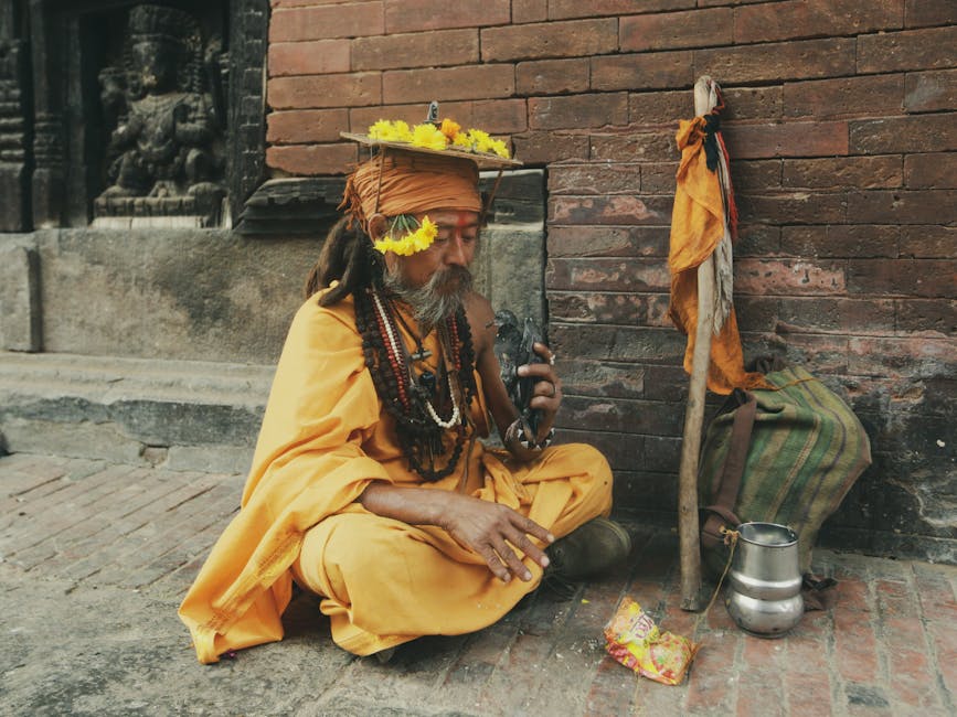A man dressed in orange robes with flower garlands sits on the ground beside a staff, bag, metal pot, and snack packet, near a brick wall with a carved statue in the background.