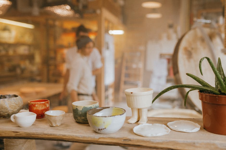 A wooden table displays various handmade ceramic bowls and a potted aloe vera plant in a pottery studio, with two people blurred in the background.