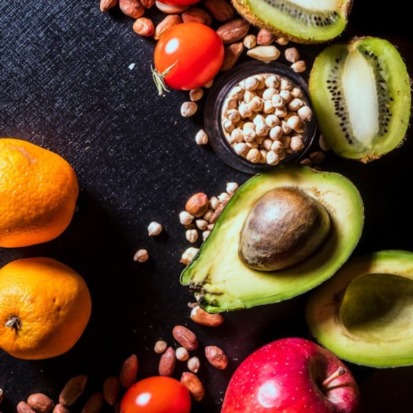 Assorted fruits and nuts, including tangerines, apple, avocado, kiwi, cherry tomatoes, peanuts, and chickpeas, arranged on a black surface.