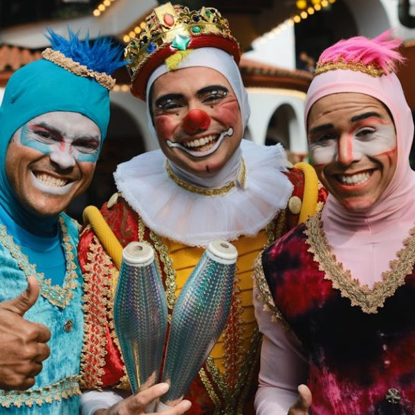 Three smiling clowns in colorful costumes and makeup pose together, with one clown holding juggling pins. Two clowns give thumbs up.