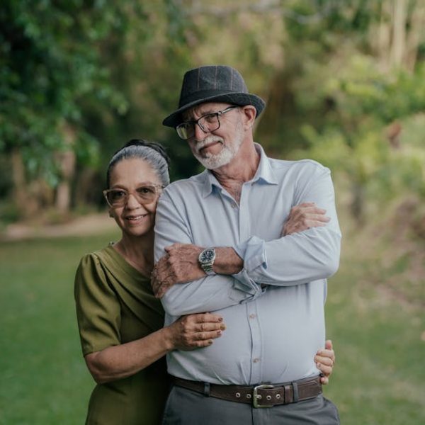 An older woman and man stand together outdoors on grass, with trees in the background. The man wears a hat and glasses; the woman is beside him, smiling and holding his arm.