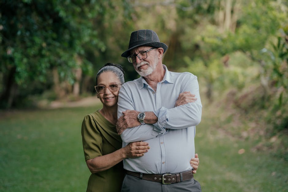 An older woman and man stand together outdoors on grass, with trees in the background. The man wears a hat and glasses; the woman is beside him, smiling and holding his arm.