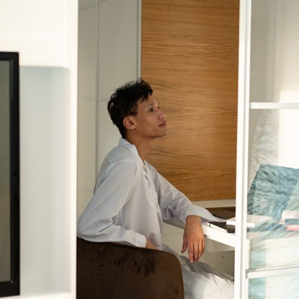 A person in a white outfit sits at a desk near a window, looking up thoughtfully. There is a laptop and stack of books on the desk.