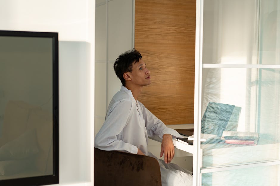 A person in a white outfit sits at a desk near a window, looking up thoughtfully. There is a laptop and stack of books on the desk.