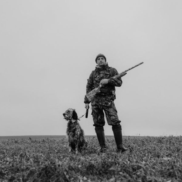 A person in camouflage clothing stands in a field holding a rifle, with a dog sitting beside them, under a cloudy sky.