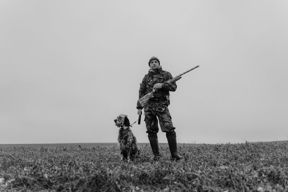A person in camouflage clothing stands in a field holding a rifle, with a dog sitting beside them, under a cloudy sky.