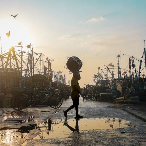 A person carrying a basket on their head walks through a wet dock area with fishing boats and equipment at sunset.