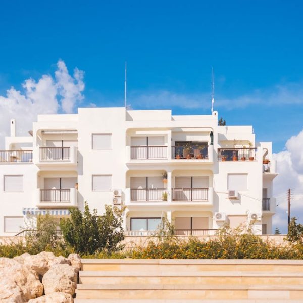 A modern white apartment building with balconies is shown under a blue sky with scattered clouds, with plants and a stone fence in the foreground.