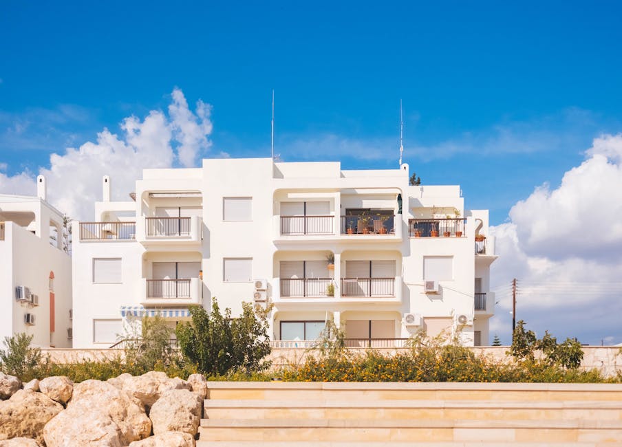 A modern white apartment building with balconies is shown under a blue sky with scattered clouds, with plants and a stone fence in the foreground.