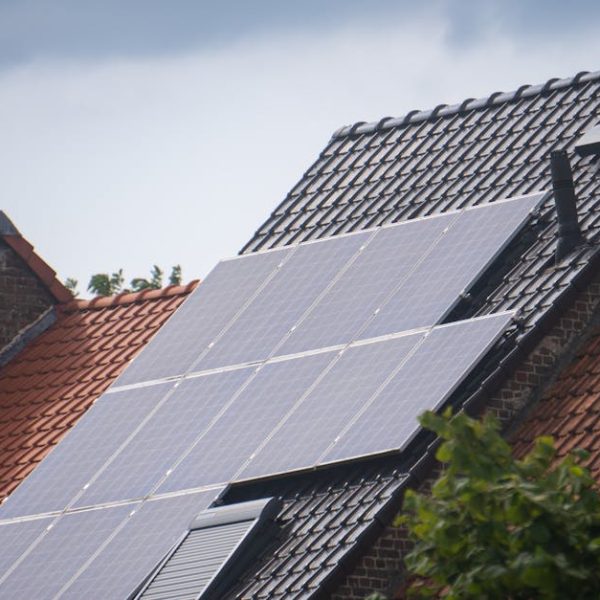 Solar panels installed on the roof of a house with dark roof tiles, adjacent to other houses with red tiled roofs, under a cloudy sky.