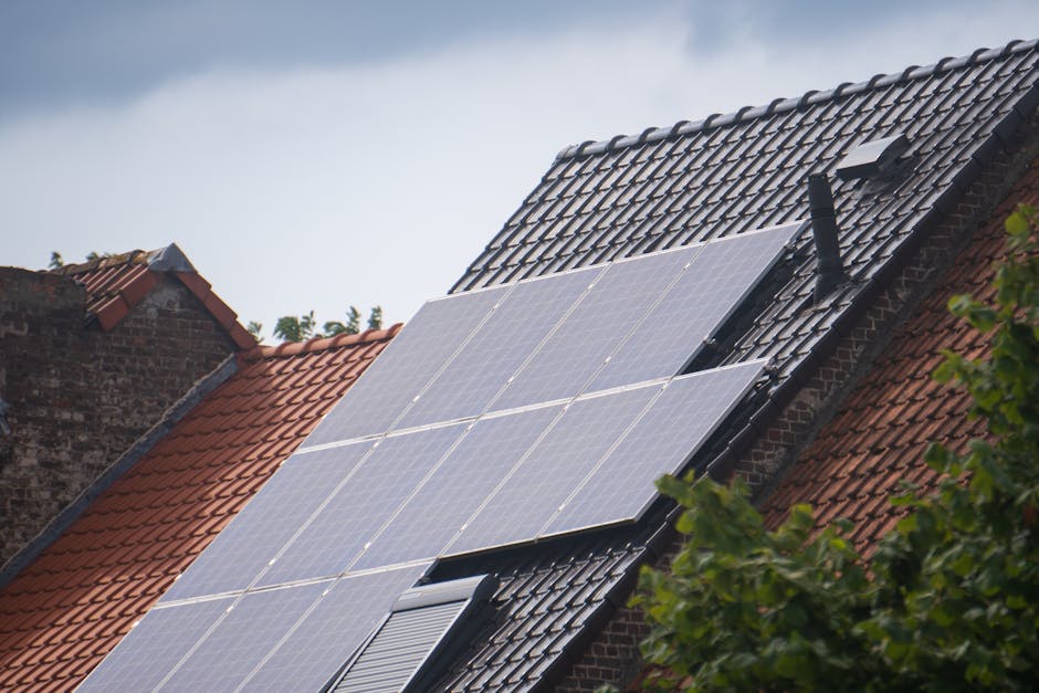 Solar panels installed on the roof of a house with dark roof tiles, adjacent to other houses with red tiled roofs, under a cloudy sky.