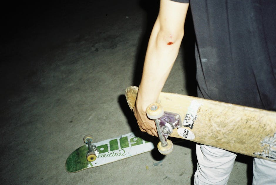 Person with a scraped elbow holds a skateboard while another skateboard lies on the ground, both on a concrete surface at night.