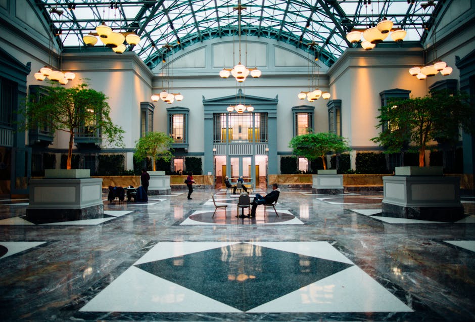 Spacious indoor atrium with a glass ceiling, marble floor, modern lighting, trees, and people sitting or walking.