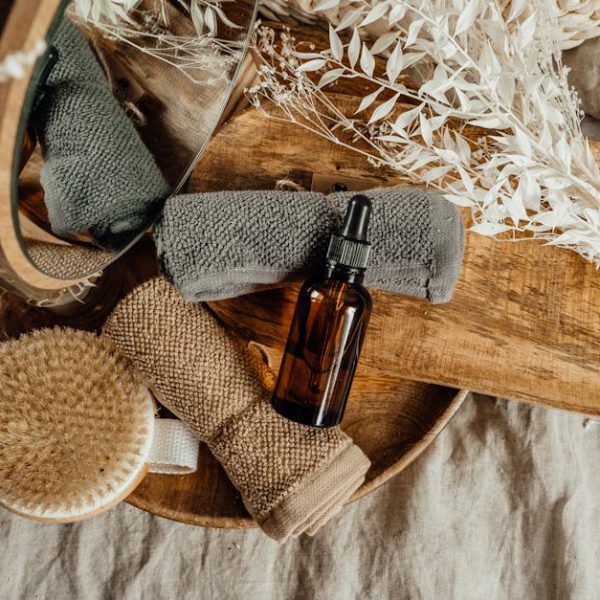 A brown glass dropper bottle, two rolled towels, a dry brush, and dried plants arranged on a wooden tray.