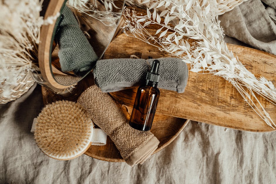 A brown glass dropper bottle, two rolled towels, a dry brush, and dried plants arranged on a wooden tray.