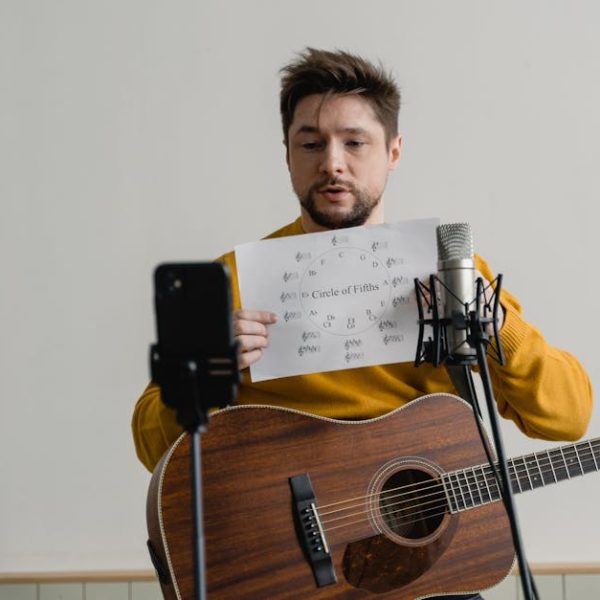 A man holding a guitar displays a sheet labeled "Circle of Fifths" in front of a microphone and smartphone on a stand.