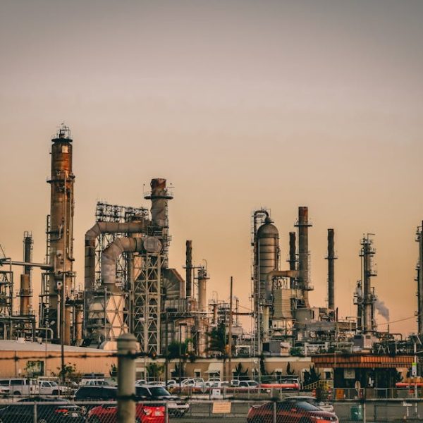 Industrial refinery complex with multiple towers and pipes, emitting light smoke, set against a sunset sky; parked cars visible in the foreground.