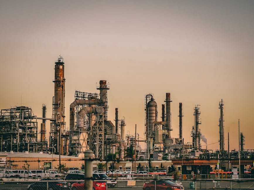 Industrial refinery complex with multiple towers and pipes, emitting light smoke, set against a sunset sky; parked cars visible in the foreground.