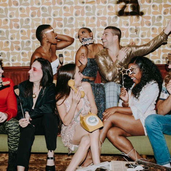A group of people in festive clothing sit on a couch, laughing and holding party props in front of a retro-patterned wall.
