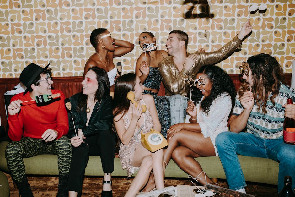 A group of people in festive clothing sit on a couch, laughing and holding party props in front of a retro-patterned wall.
