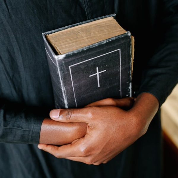 Person in a black robe holding a closed Bible with a cross on the cover, arms crossed over the book.