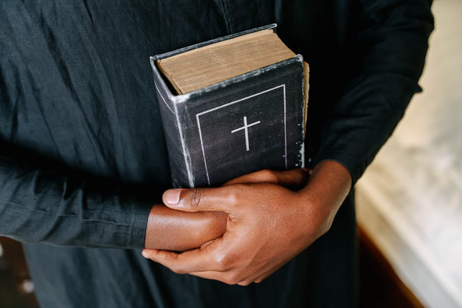 Person in a black robe holding a closed Bible with a cross on the cover, arms crossed over the book.