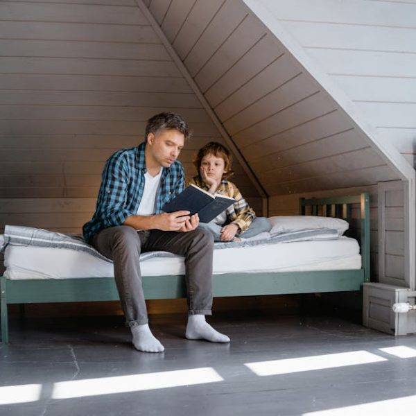 An adult and a child sit on a bed in a minimalist attic room, with the adult reading from a book while the child listens attentively.
