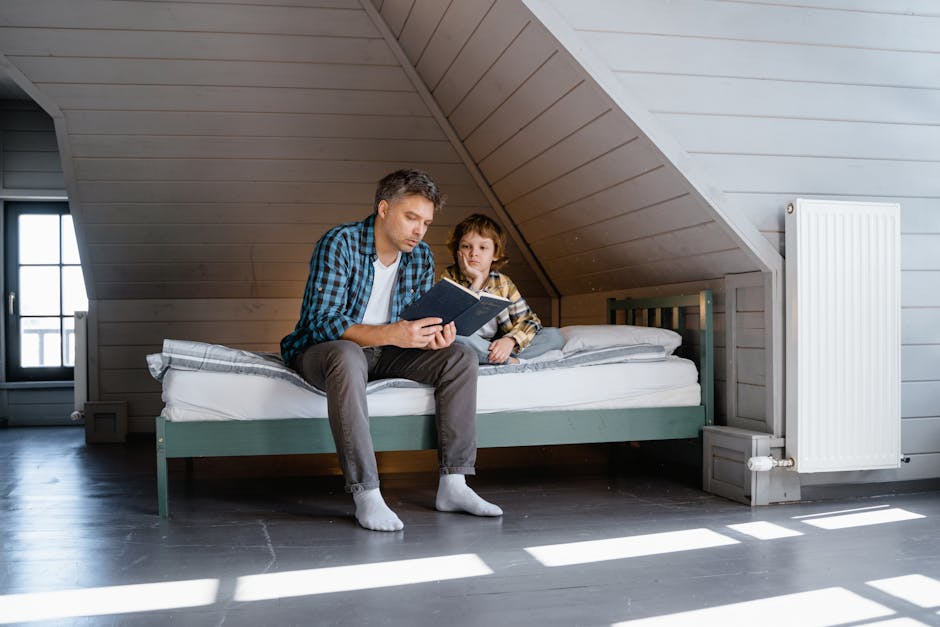 An adult and a child sit on a bed in a minimalist attic room, with the adult reading from a book while the child listens attentively.