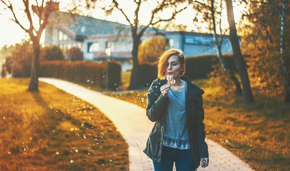 A person stands on a sunlit path in a park, blowing seeds from a dandelion. Trees and a house are visible in the background.