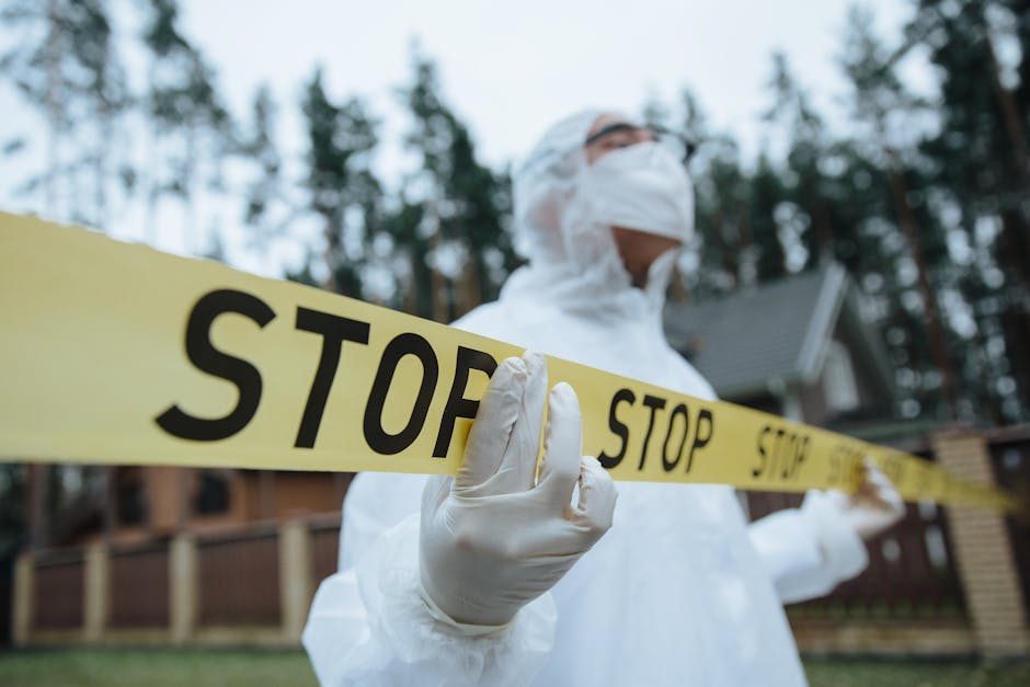 Person in protective suit and mask holding yellow tape marked "STOP" outdoors near houses and trees, suggesting a restricted or hazardous area.