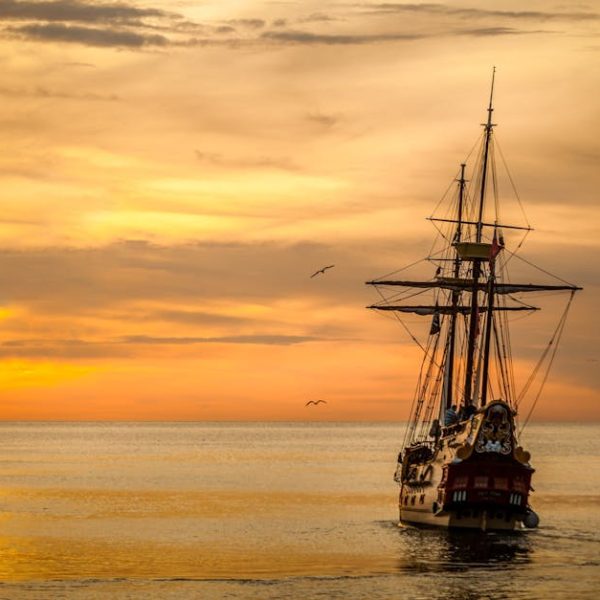 A sailing ship with tall masts moves across calm water at sunset, under a golden sky with a few birds flying nearby.
