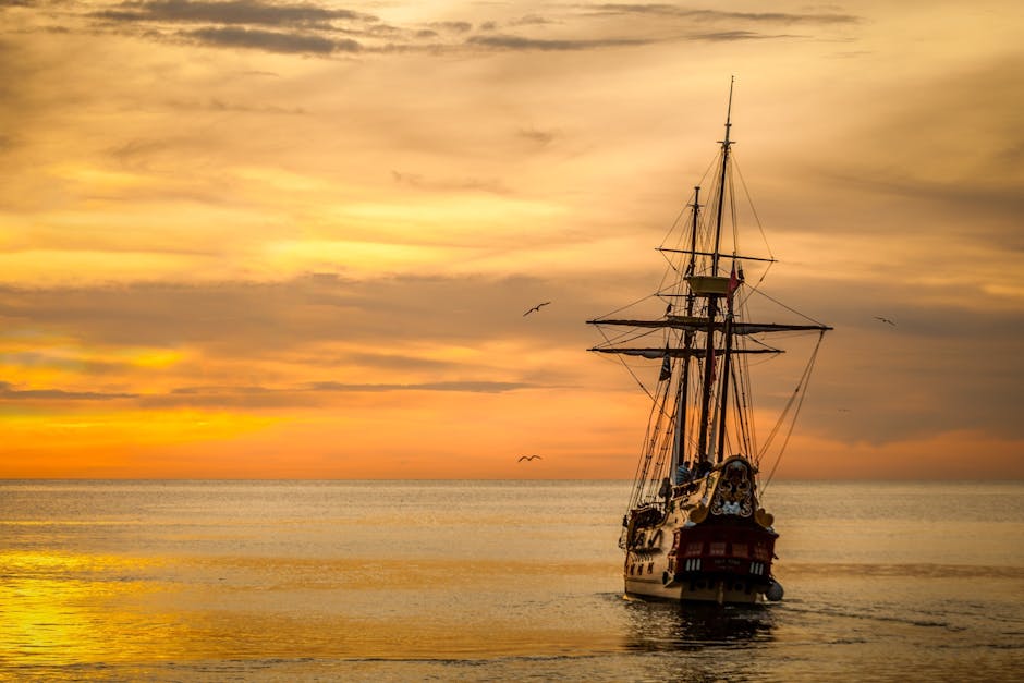 A sailing ship with tall masts moves across calm water at sunset, under a golden sky with a few birds flying nearby.