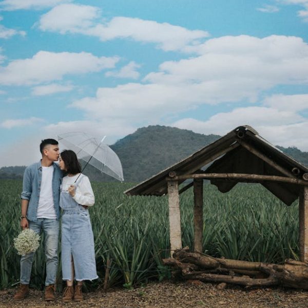 A couple stands in front of a field, with one person kissing the other on the forehead while holding an umbrella and flowers, next to a small wooden shelter under a blue sky with clouds.