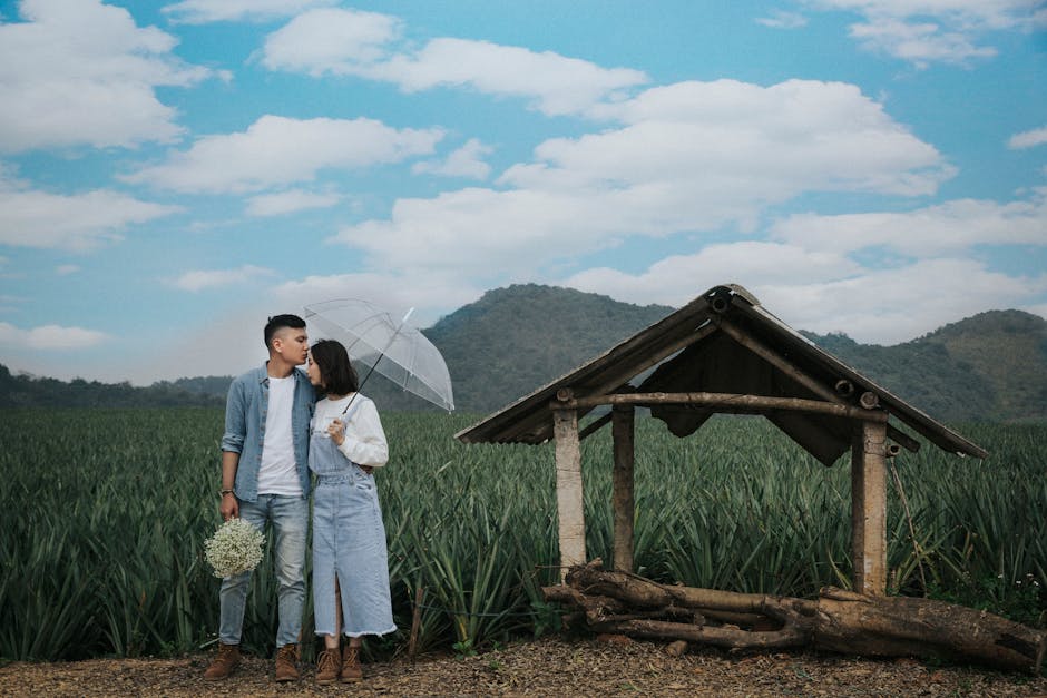 A couple stands in front of a field, with one person kissing the other on the forehead while holding an umbrella and flowers, next to a small wooden shelter under a blue sky with clouds.