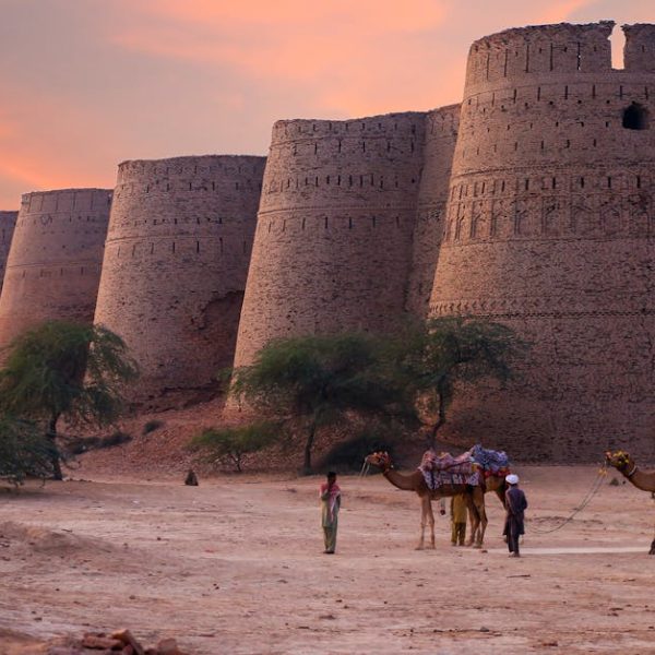 Two people with camels stand near the large, ancient brick walls of a fort at sunset, with sparse trees and sandy ground in the foreground.