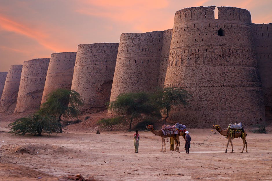 Two people with camels stand near the large, ancient brick walls of a fort at sunset, with sparse trees and sandy ground in the foreground.