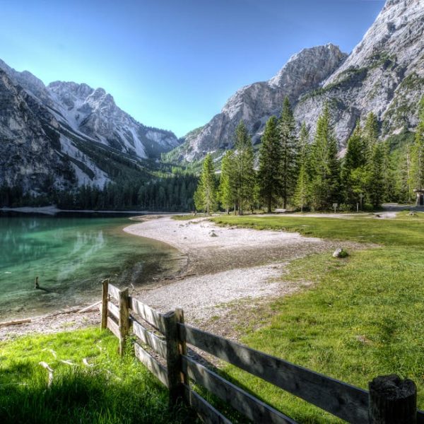 A clear lake is surrounded by mountains, pine trees, and green grass, with a wooden fence and a gravel shore in the foreground under a blue sky.