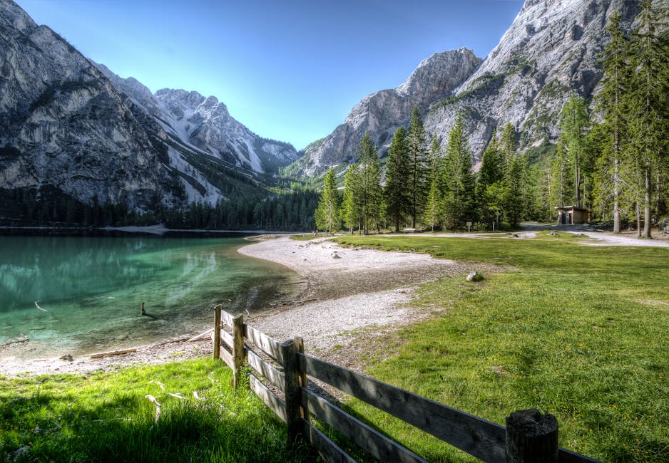 A clear lake is surrounded by mountains, pine trees, and green grass, with a wooden fence and a gravel shore in the foreground under a blue sky.