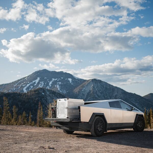 A silver angular vehicle is parked on a dirt road with a mountain range and scattered clouds in the background.