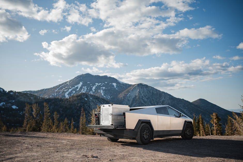 A silver angular vehicle is parked on a dirt road with a mountain range and scattered clouds in the background.