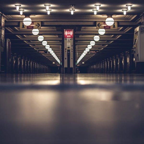 Dimly lit underground hallway with rows of round ceiling lights, reflective floors, and a red emergency SOS sign in the center.