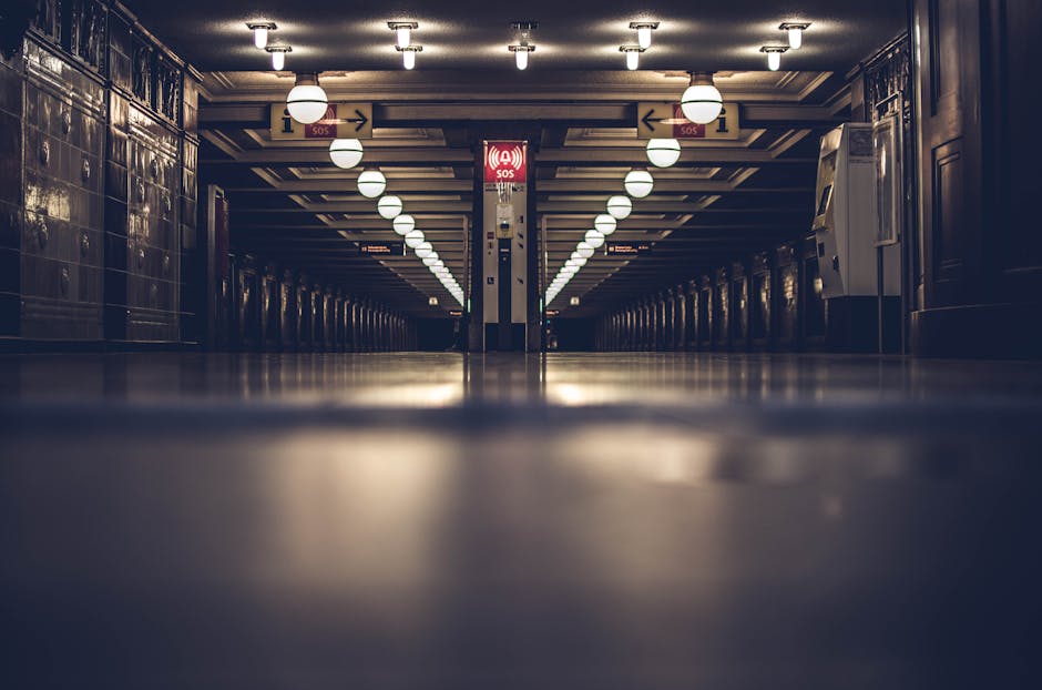 Dimly lit underground hallway with rows of round ceiling lights, reflective floors, and a red emergency SOS sign in the center.