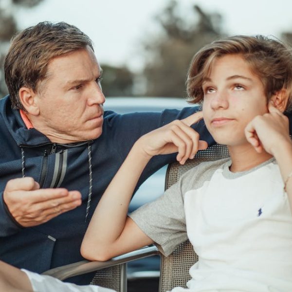 An adult sits beside a teenager, gesturing with his hand while the teenager looks away and covers their ears with their fingers.