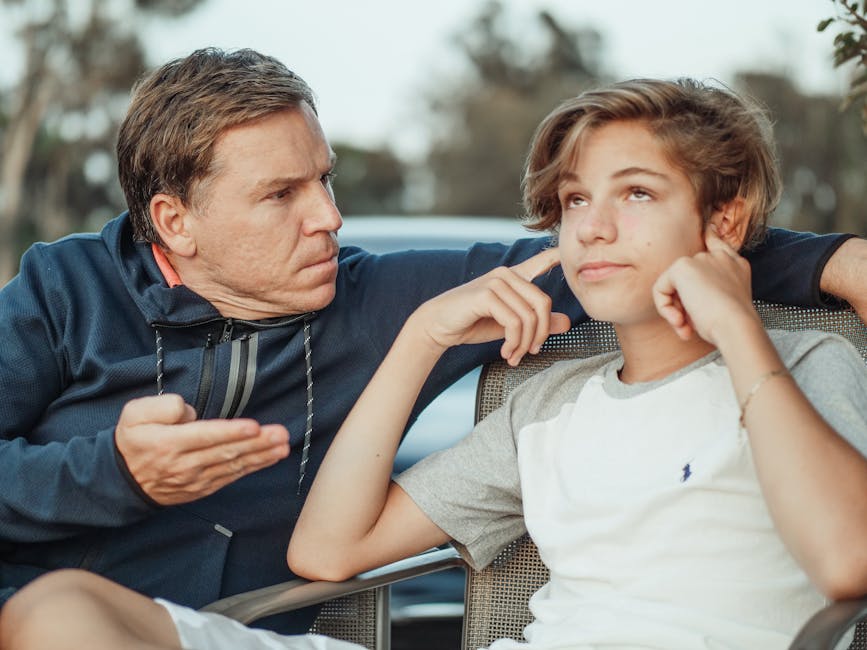 An adult sits beside a teenager, gesturing with his hand while the teenager looks away and covers their ears with their fingers.