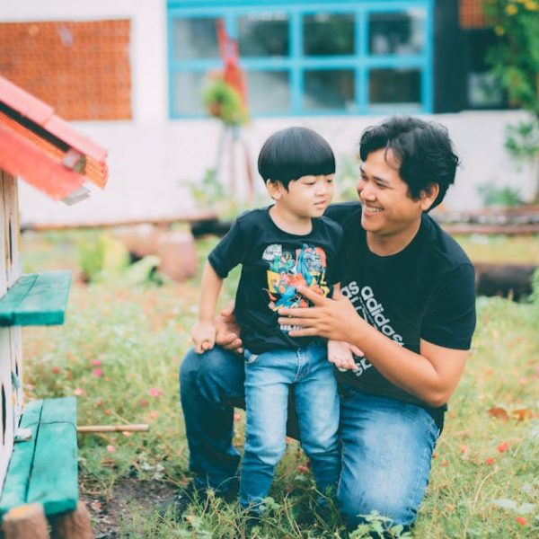 An adult and a child kneel together outside by a green structure, both wearing black t-shirts and jeans, with greenery and a building in the background.