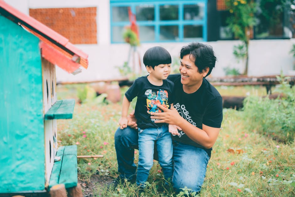 An adult and a child kneel together outside by a green structure, both wearing black t-shirts and jeans, with greenery and a building in the background.